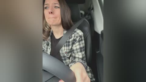 A video of a young woman with shoulder-length brown hair, wearing a black shirt under a plaid shirt, driving in a car, with a neutral expression, blurred background.