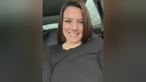 Video of a smiling woman with straight brown hair, wearing a black long-sleeve top, sitting in a car with the window down, blurred background.