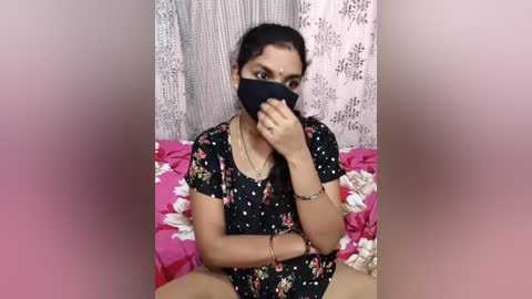 Video of a young South Asian woman with medium skin tone, wearing a black floral dress, black face mask, and a silver bracelet. She sits on a bed with pink and white floral sheets, against a backdrop of white and beige lace curtains.