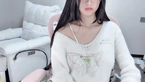 Video of an Asian woman with long black hair, wearing a white knit sweater with a pink chair and white leather couch in the background.