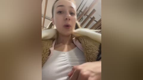 A video of a blonde woman in a white tank top and beige fur-trimmed jacket, with a surprised expression, taken from a close-up angle. Background shows a blurred indoor setting with wooden shelves.