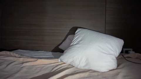 Video of a dimly lit bedroom with a white pillow on a beige bedspread. The background shows a wooden-paneled wall and an electrical outlet, suggesting a minimalist, modern decor.
