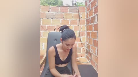 Video of a fit woman with medium skin tone, dark hair in a bun, wearing a black sports bra and pants, sitting on a black exercise mat in a small, brick-walled outdoor gym area.