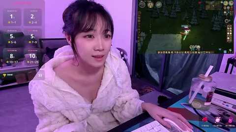 Video of an Asian woman with black hair in a white bathrobe, seated at a gaming desk, playing a video game with an Asian setting, lit by purple and green light.