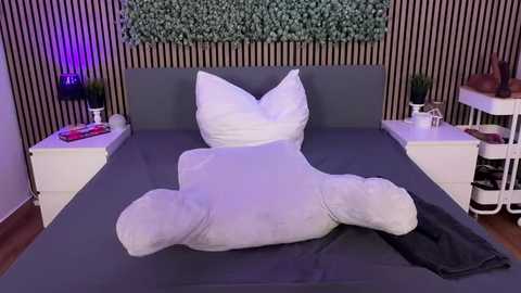 Video of a modern bedroom with a gray bed, white pillows, and a plush white teddy bear. Black-and-white striped wallpaper, white nightstands with lamps, and a green leafy wall decoration.