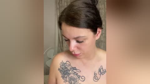 Video of a young woman with fair skin and dark hair, adorned with a floral and cursive tattoo on her chest. She gazes downward, wearing minimal makeup, in a room with beige walls and a fan visible in the background.