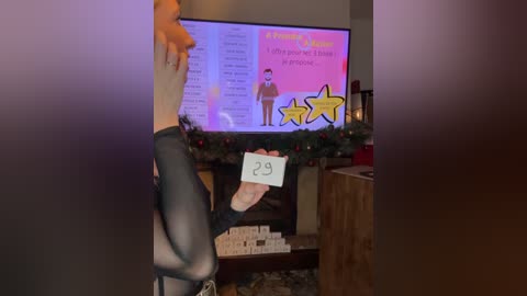 Video of a woman holding a sign with a 29 written on it, standing in front of a TV showing a \"Bachelor\" episode, decorated with holiday lights and a wreath.