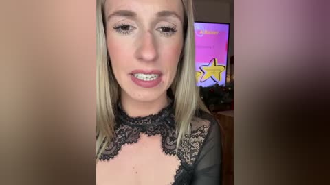 A video of a blonde woman with fair skin and braces, wearing a black lace top, standing in a room with a TV displaying a star symbol and Christmas decorations in the background.