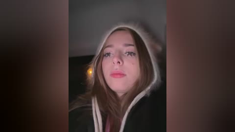 Video of a young woman with light skin, brown hair, and a neutral expression, wearing a black and white hooded jacket, standing in a dimly lit room.