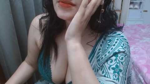 A video of a young woman with fair skin and long black hair, wearing a teal lace bra that reveals cleavage, lying on a pink floral bed. She is touching her face with her left hand.