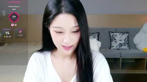 A video of an Asian woman with long black hair, wearing a white top, smiling in a modern, minimalist living room with a gray sofa and black decorative pillows.