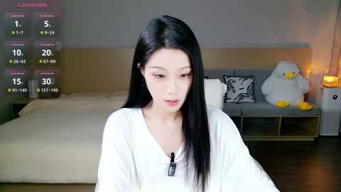 Video of an East Asian woman with long black hair, wearing a white shirt, in a modern bedroom with a beige bed, white pillow, and stuffed toys.
