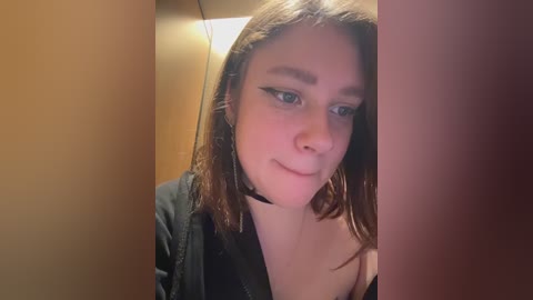 A video of a young woman with fair skin, brown hair, and a black choker, standing in a dimly lit room with beige walls, wearing a black top.