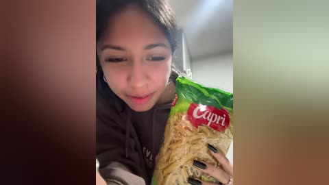 Video of a young woman with medium brown skin, dark hair, and black nails, wearing a dark hoodie, holding a green and red Cappi pasta packet, smiling.