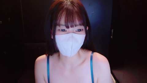 Video of an East Asian woman with straight, shoulder-length dark hair and pale skin wearing a white surgical mask and a blue spaghetti-strap top, set against a dimly lit, dark background.