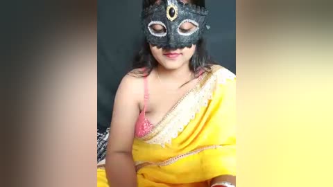 A video of a South Asian woman in a bright yellow sari with a lace border, wearing a black Venetian mask with intricate lace patterns and a pink bra. The background is blurred.