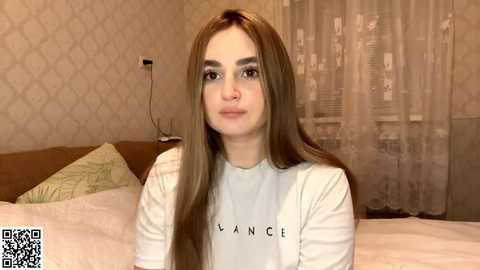 Video of a young woman with long brown hair, light skin, and blue eyes, wearing a white shirt with \"Bianca\" in black letters, sitting on a bed with beige wallpaper and white curtains in the background.