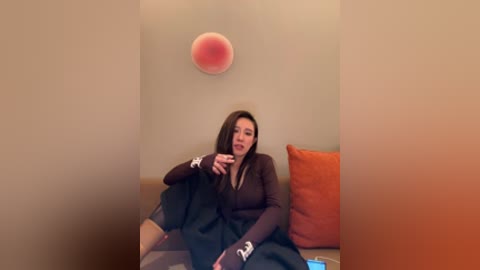 Video of a woman with light skin and straight brown hair, wearing a brown cardigan and black skirt, sitting on a beige couch, playing with a red stress ball, orange cushion beside her.