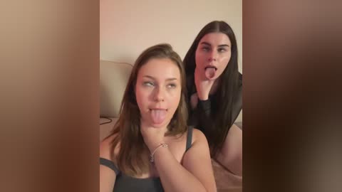 Video: Two young women, one with brown hair, another with black hair, playfully stick out their tongues. They wear casual tops, seated on a bed with beige pillows.