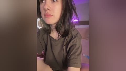 Video of a young woman with straight, dark hair, wearing a dark T-shirt, looking thoughtful. Background includes a bed with pink sheets, a circular light, and a purple light source.