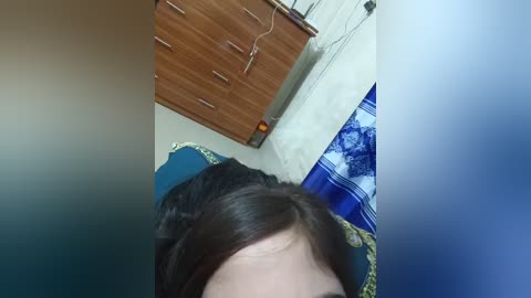 A video showing a close-up of a person's face, light skin, and brown hair, lying on a bed with a blue and yellow patterned blanket, in a room with a wooden dresser and blue curtains.