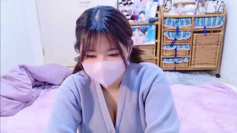 A video of a young Asian woman with brown hair in a wicker basket bed, wearing a white surgical mask and a light blue robe, in a cozy bedroom with a white door and wicker storage unit.
