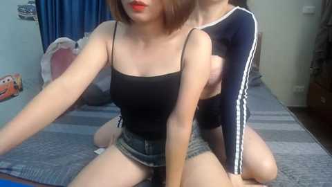 Video of two women in a bedroom: one in a black tank top, denim shorts, and red lipstick; the other in a navy blue long-sleeve top. They are sitting on a striped bedspread.