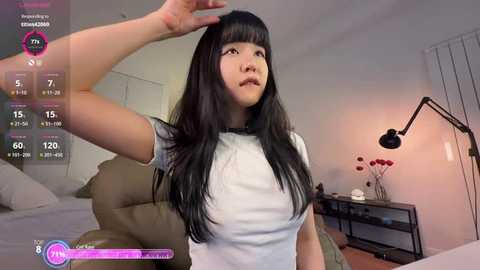 Video of an East Asian woman with long black hair, wearing a white t-shirt, adjusting her hair in a modern living room with a beige couch, black floor lamp, and digital screen displaying live stream details.
