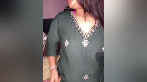 A video shows a woman with medium brown skin, wearing a dark green, long-sleeved blouse with intricate silver embroidery and a deep V-neck, standing indoors with blurred background.