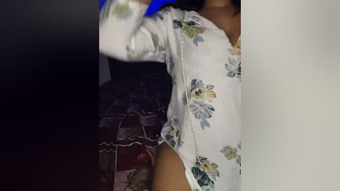 Video of a woman with medium brown skin wearing a floral-patterned white blouse and a maroon skirt with lace detailing. She has a slender physique, and her face is not visible. The background is dark and blurry.