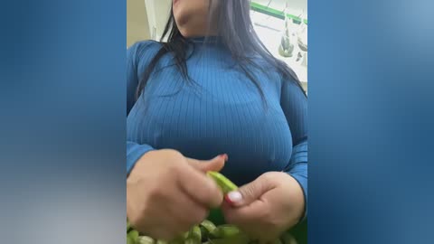 Video of a woman with long black hair, wearing a tight, blue ribbed long-sleeve top, holding a green pepper, indoors with a blurred background.