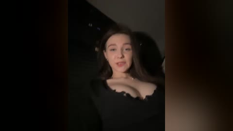 Video of a young woman with fair skin, dark hair, and a black dress with a plunging neckline, standing in a dimly lit room, with blurred edges.