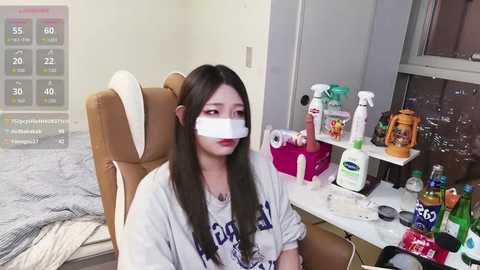 A video of a young woman with long dark hair, wearing a white T-shirt with a black logo, sitting on a beige chair, with a white bandage covering her mouth. The room has a cluttered desk with various items, including alcohol bottles and cleaning products.