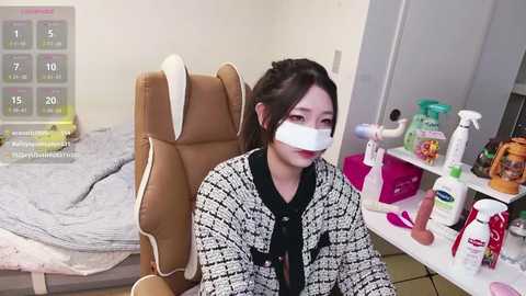 Video of a young woman with black hair, wearing a black and white checkered coat, sitting in a beige recliner chair with a white mask covering her nose and mouth. The background features a cluttered desk with cleaning supplies, a bed with a gray blanket, and a calendar.