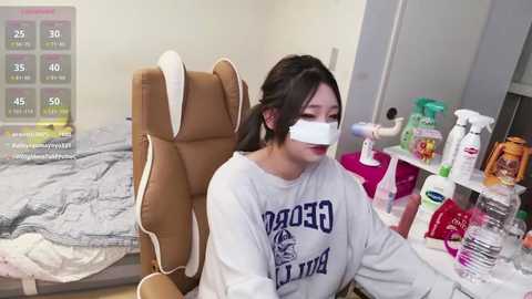 Video of an Asian woman in a white sweatshirt, wearing a white face mask, sitting in a tan gaming chair, surrounded by cleaning supplies and a cluttered desk.
