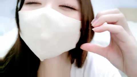Video of a young Asian woman with long brown hair, wearing a white surgical mask, closed eyes, and a white top, smiling.