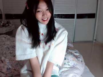 Video of an Asian woman with long black hair, wearing a white sweater, sitting on a bed with floral bedding, smiling in a cozy bedroom with white blinds.