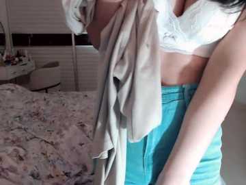 Video of a woman in a white lace bra and turquoise pants, holding a beige towel, standing next to a messy bed with floral sheets in a dimly lit room with white blinds.