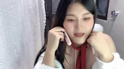 Video of an East Asian woman with long black hair, wearing a white off-shoulder dress, holding a pink earpiece, looking into a mirror in a bathroom with textured white walls and a window.