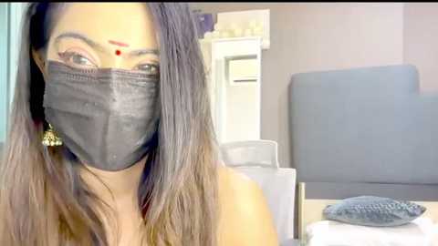 Video of a woman with long, dark hair and a black mask, wearing a red dot on her forehead, standing in a medical room with a gray chair and white cabinet.