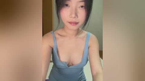 Video of a young Asian woman with light skin and dark hair, wearing a low-cut blue tank top, posing indoors with a neutral background.