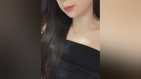 Video of a light-skinned woman with long, wavy black hair, wearing a black off-shoulder top, and a delicate necklace. Her face is partially visible, showing full lips and a small silver pendant. The background is a gradient of dark to light brown.