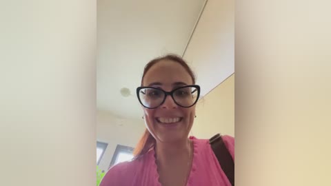 A video of a smiling woman with glasses, wearing a pink top, standing indoors with beige walls and a window visible in the background.