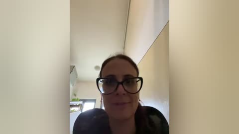 Video of a smiling woman with light skin and brown hair, wearing black-rimmed glasses and a dark jacket, standing in a narrow, sunlit hallway with beige walls.