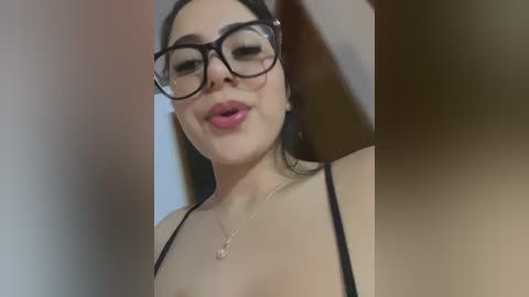 Video of a woman with fair skin, dark hair, and large glasses, wearing a black spaghetti strap top, smiling with closed lips. Background is blurred.