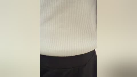Video of a person's torso, showing a white ribbed sweater over black pants, against a plain beige wall background.