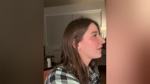 Video of a young woman with light skin and long brown hair, wearing a checkered shirt, looking sideways in a dimly lit room with a beige wall and a wooden table.