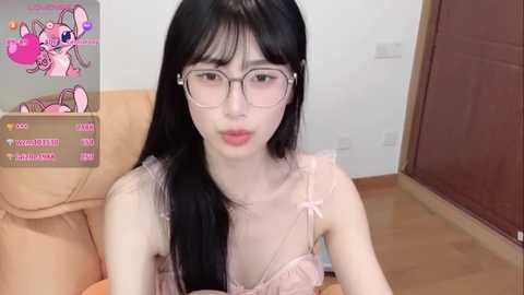 A video of an East Asian woman with long black hair, wearing glasses and a pink lace bra, sitting on a beige couch in a simple room.