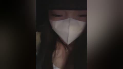 A dimly-lit video of a person wearing a white medical mask, partially obscured by a dark, shadowy background, suggesting a somber or secretive atmosphere.