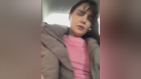 A blurred, candid video shows a woman in a car, wearing a grey puffer jacket over a pink shirt, with her head tilted back, eyes closed, seemingly asleep or in deep thought.
